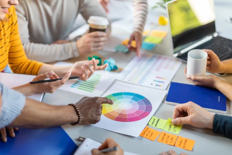 A team around a desk covered in colour wheels, printed colour charts and sticky notes, one hand pointing at a colour wheel